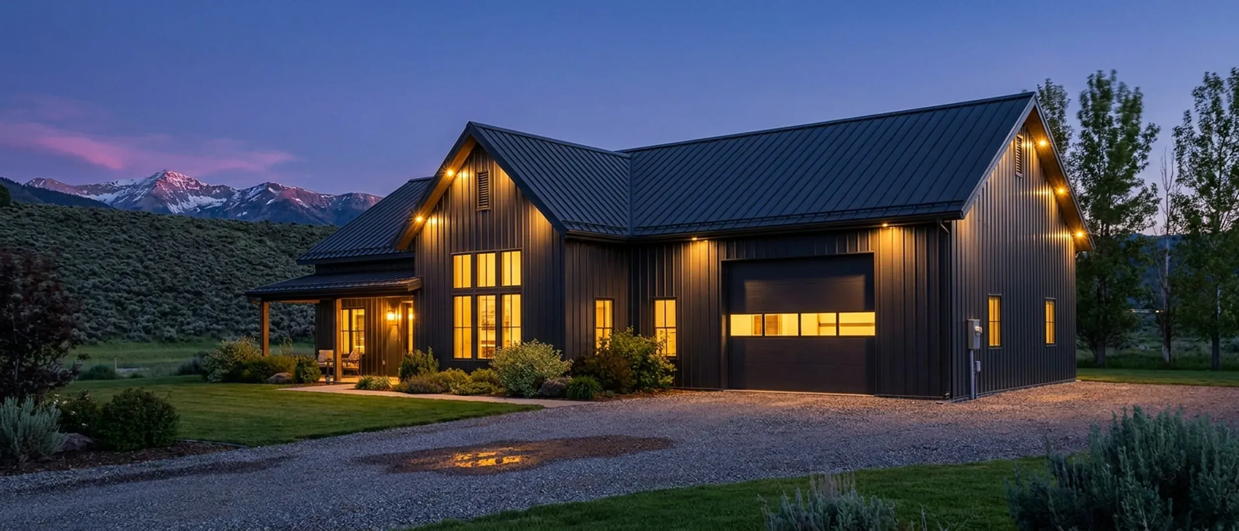 A barndominium at dusk with warm light glowing from every window, sagebrush foothills and a snow-capped mountain range behind it against a deep blue twilight sky.