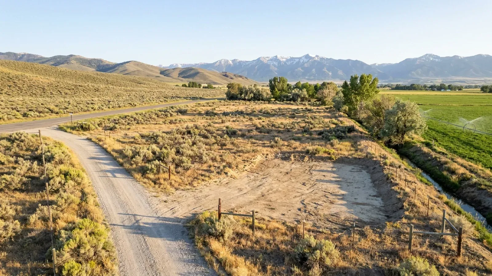 Aerial view of an undeveloped rural lot of dry pasture and sagebrush with a gravel access track, an irrigation ditch lined with cottonwoods and green hay fields beyond.